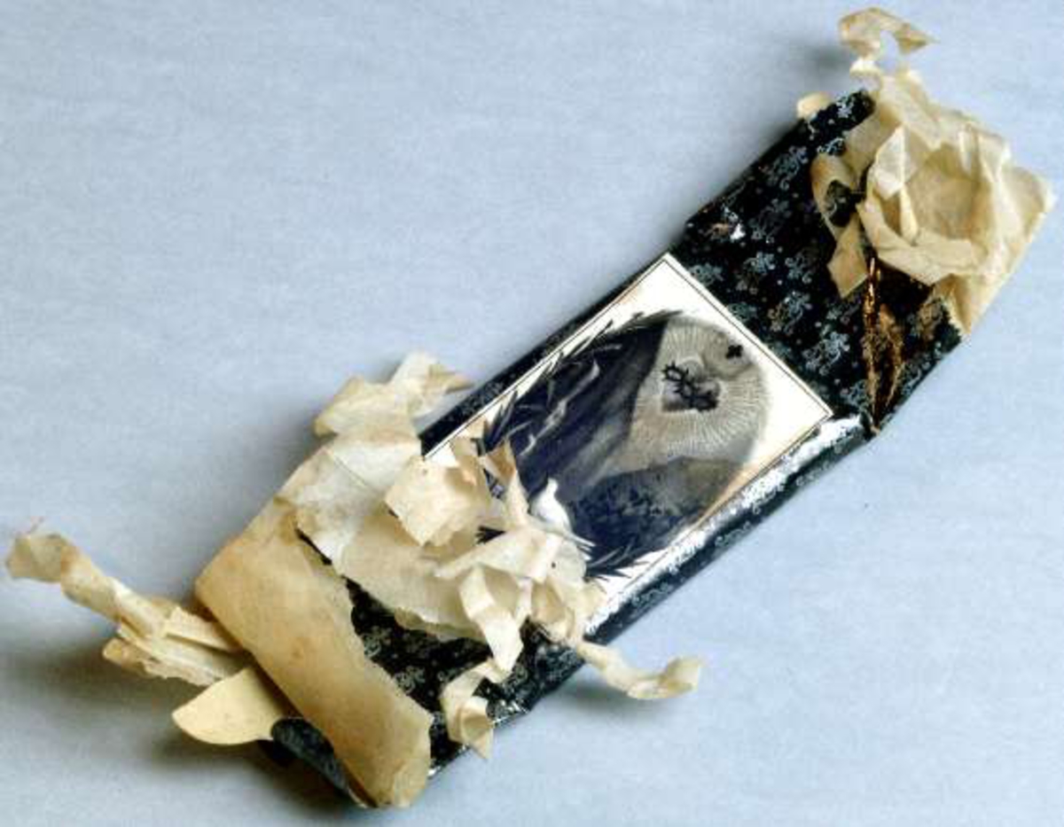 Begravningskaramell med omslag av beige silkespapper och svart glanspapper med tryckta silverfärgade medaljonger. Pappersetikett med tryck symboliserande treenigheten.