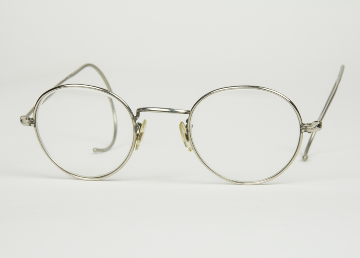 Runda glasögon med bågar av metall. Rektangulärt glasögonfodral av grå metall. I fodralet ligger även duk för rengöring av glasögonen.
