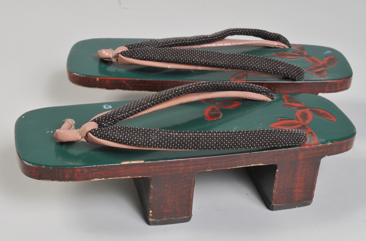 Sandaler i tre (japansk fottøy bestående av treplate med to trestykker på undersiden og skinnrem oppå). Brunbeiset treverk med grønnmålt overside , i oversiden enkel utskjæring av rødmalte blader.