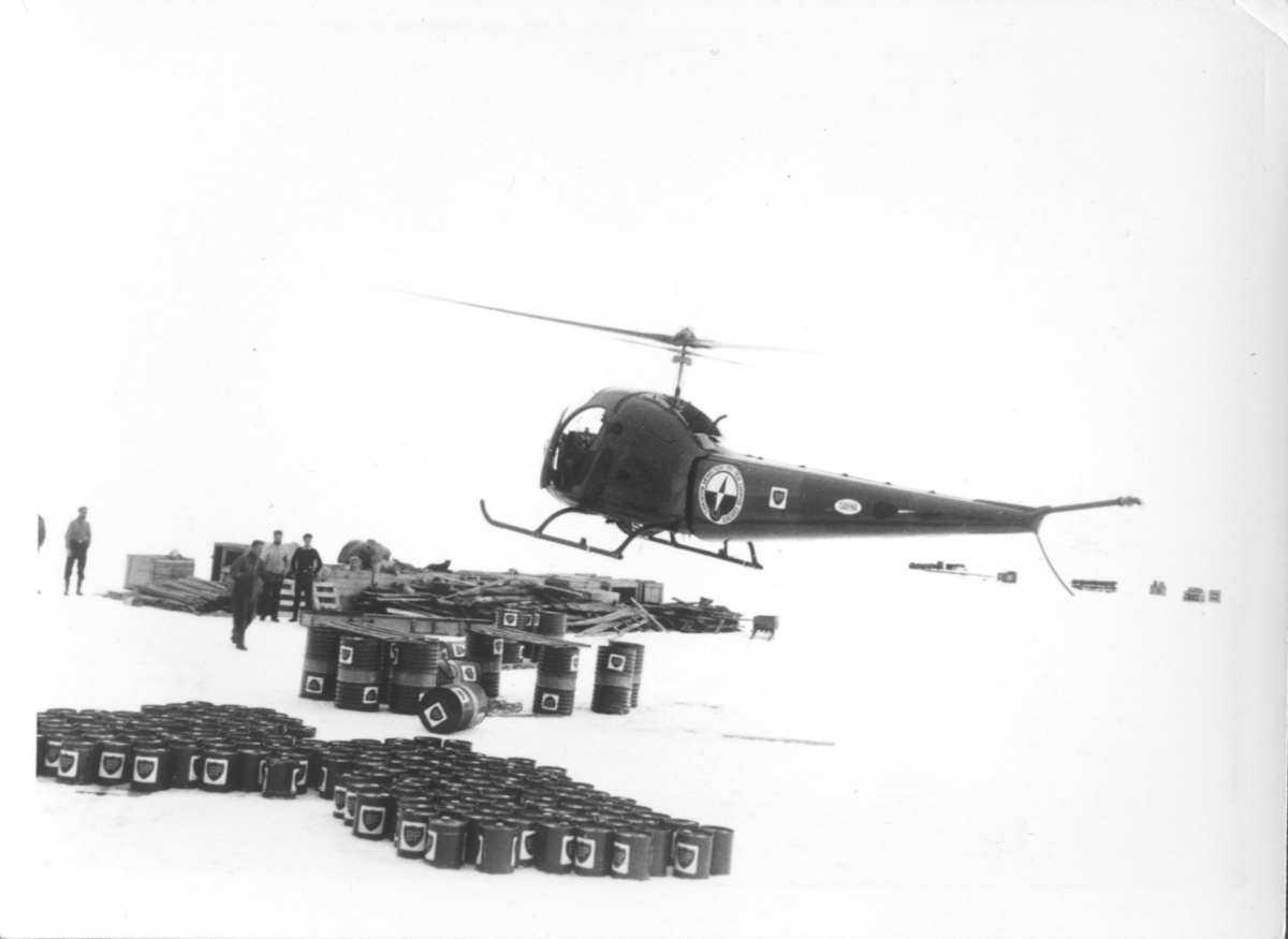 Helikopter, muligens et Bell 47H, frakter bensin frem til bensinfatene som er losset på isen. Antarktis (Sydpolen), 1957.