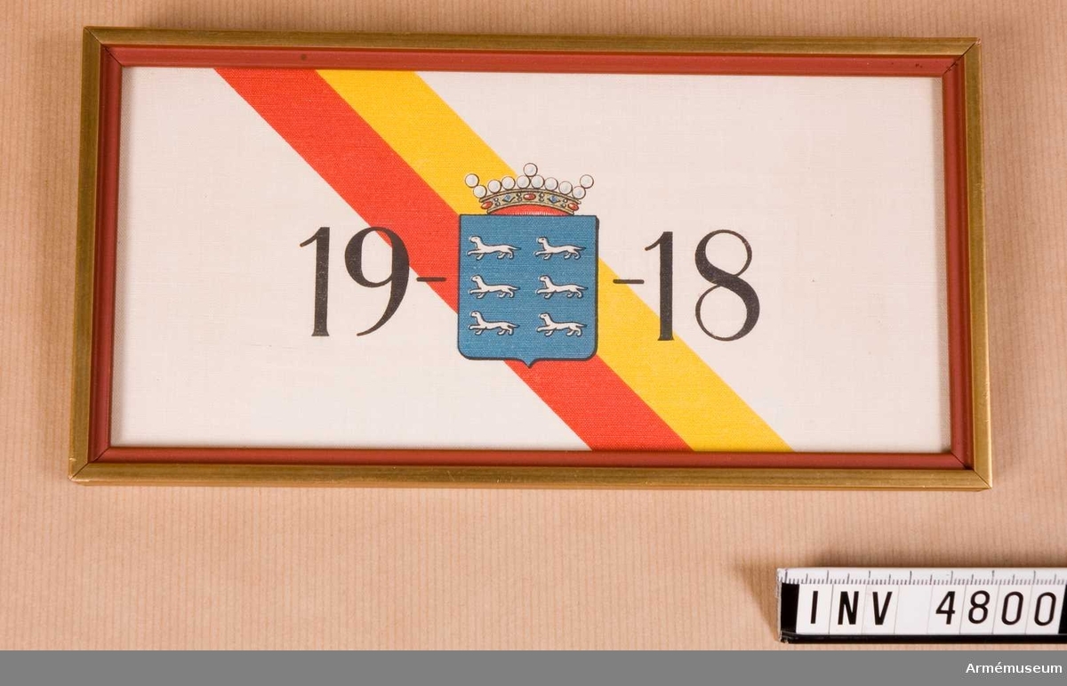 Inramad och inglasad minnestavla med Österbottens vapen och årtalet 1918 i tryck på vit väv av bomull eller linne.