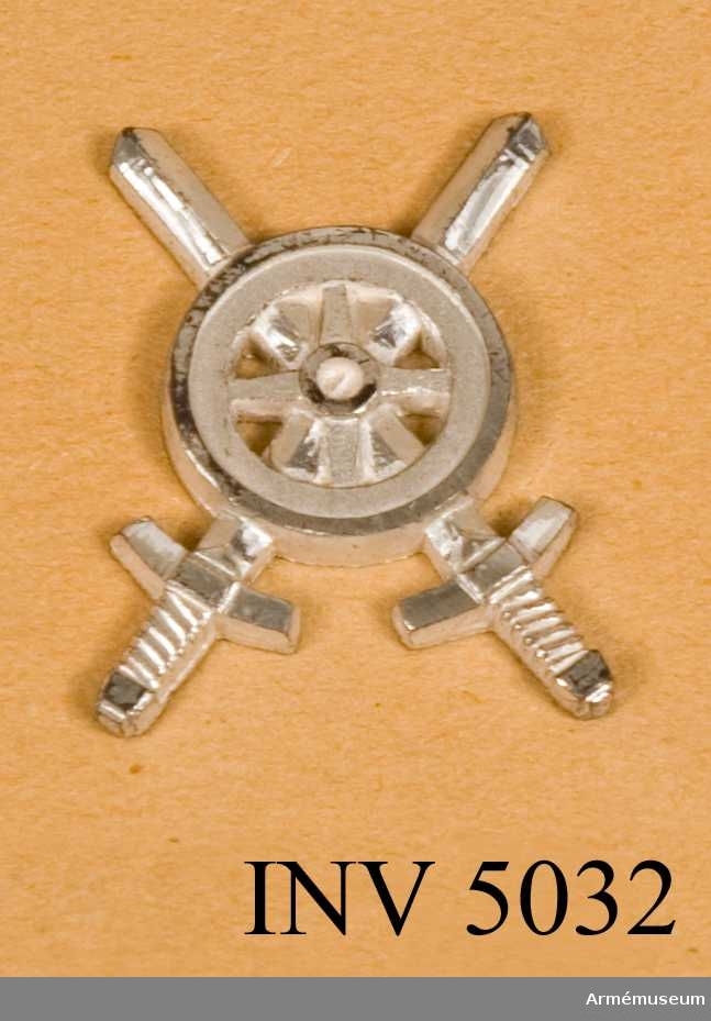 Tillverkat av metall. I likhet med emblemen till  trängtruppernas äldre uniformer är det här utfört i silverfärg. Samma motiv som i 1939 års modell, ett kärrhjul på två korslagda svärd, men något annorlunda utformat.
