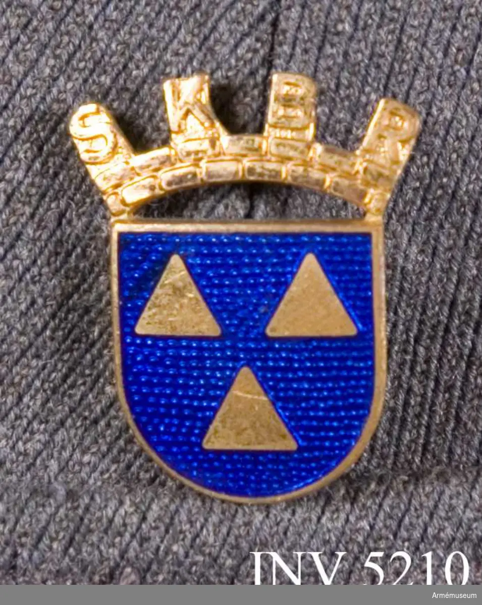 Brosch, SKBR. 1950. Höjd: 30 mm. Av förgyllt mässing och blå emalj. Bäres på jackans vänstra strax ovan ficklocket.