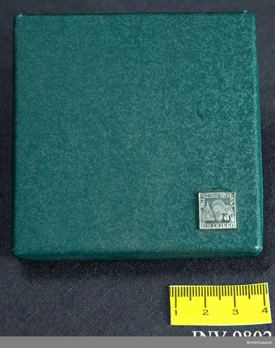 Ask av grön klot tillhörande AM.009801. På lockets nedre högra hörn är fästad en miniatyr av inneliggande minnesplakett. Insidan är klädd med svart sammet och har i botten en för plaketten avpassad nedsänkning.