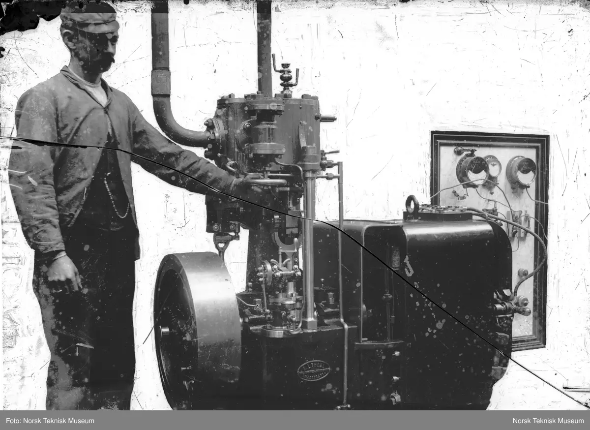 Mann som skrur på dampmaskin merket A.L. Thune Mek. Værksted & Jernstøberi Christiania. Maskinen er laget på Thunes Mekaniske Verksted på Skøyen i Oslo. Bakgrunn er dekket over med maling.