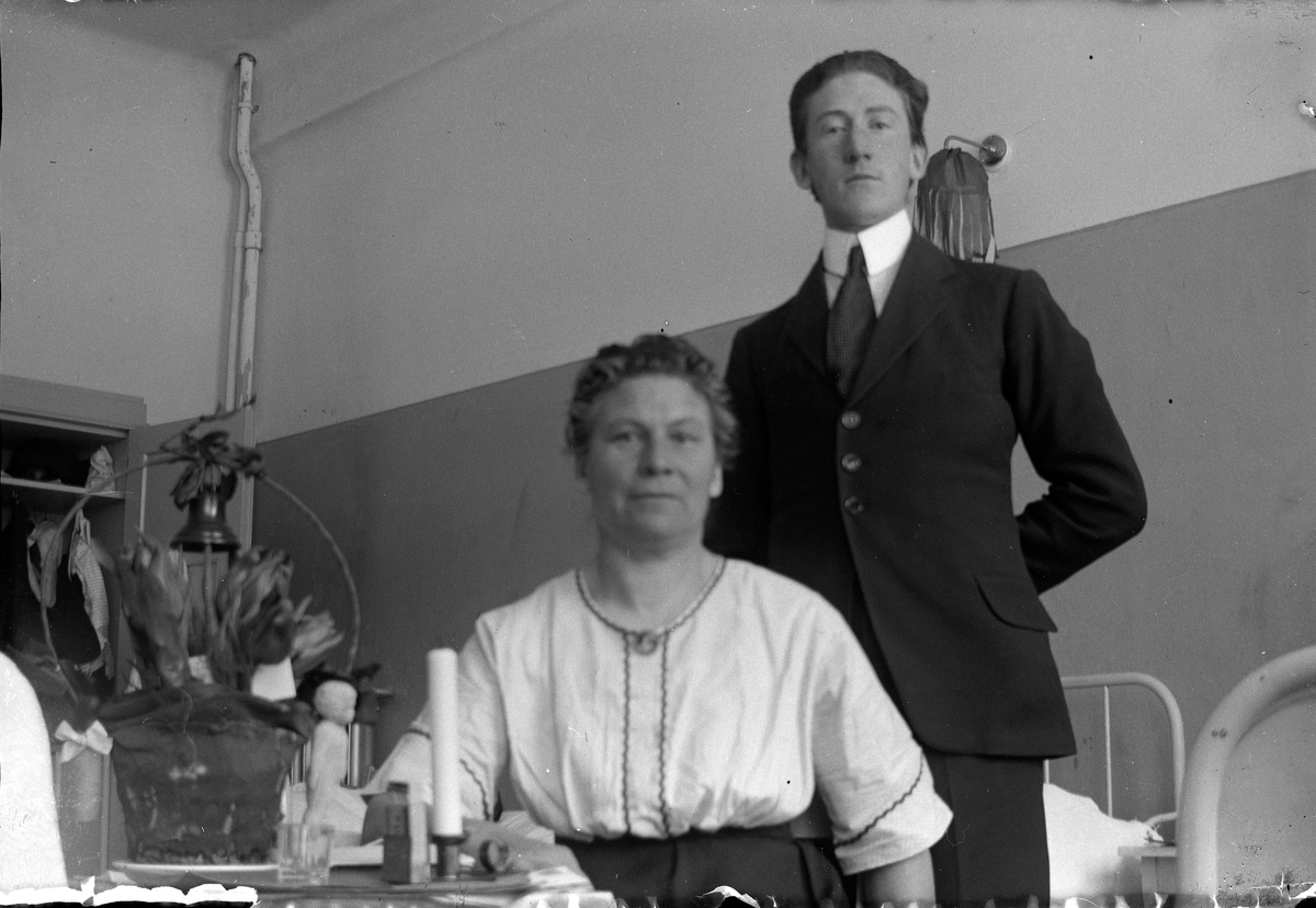 En stående man samt en kvinna sittande vid ett bord prydd med en blomsterkorg, i bakgrunden sjukhusängar, Lungkliniken, Eksjö.