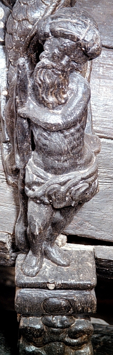 Skulptur av en naken man, så kallad vildman, stående på hög sockel.

Bålen är frontalställd medan huvudet är kraftigt lutat åt vänster och något vridet. Vildmannen har långt skägg och en virad turban på hjässan. Kring livet bär han en gördel sammansatt av växtdelar. Utmed höger sida håller vildmannen med bägge händer en lång klubba med förtjockad nederända. Kroppstyngden vilar helt på vänster ben och höft, sistnämnda markant utskjuten. Högerbenet är böjt och fötterna placerade tätt intill varandra. Figuren står inte mitt på ståplattan utan är förskjuten något åt sidan.



Sockeln är horisontell upptill och snedskuren nedtill, med en grotesk maskaron under en rektangulär vulst med utbuktande ändar. Figuren är rundskulpterad medan sockelns baksida är plan. Basen är horisontal.



Skulpturen är mycket välbevarad.



Text in English: Sculpture of a half-naked man standing on a high pedestal.

The torso is shown en face whilst the head is sharply bowed and turned to the left. The figure has a long beard, wears a turban on his head and a girdle of vegetation around the waist. With both hands he holds a long club, which widens at the base, at his right side. The weight of his body rests wholly on his left leg, his left hip is pronounced. The right leg is bent, the feet are close together. The figure does not stand in the centre of the pedestal but is placed a little to the side.



The pedestal has a rectangular horizontal entablement and a grotesque mask beneath the beading. While the figure is sculpted in the round, the back of the pedestal is flat.



The sculpture is very well preserved.