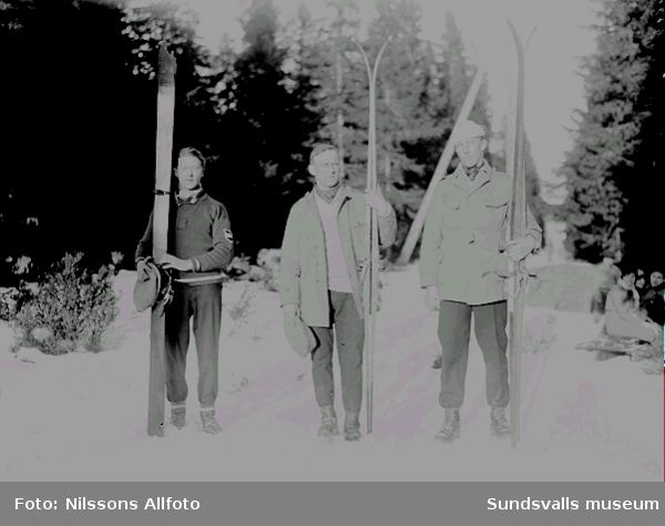 Tre män som står i skidspåret och visar upp sina skidor. Människor sitter bredvid skidspåret i bakgrunden.