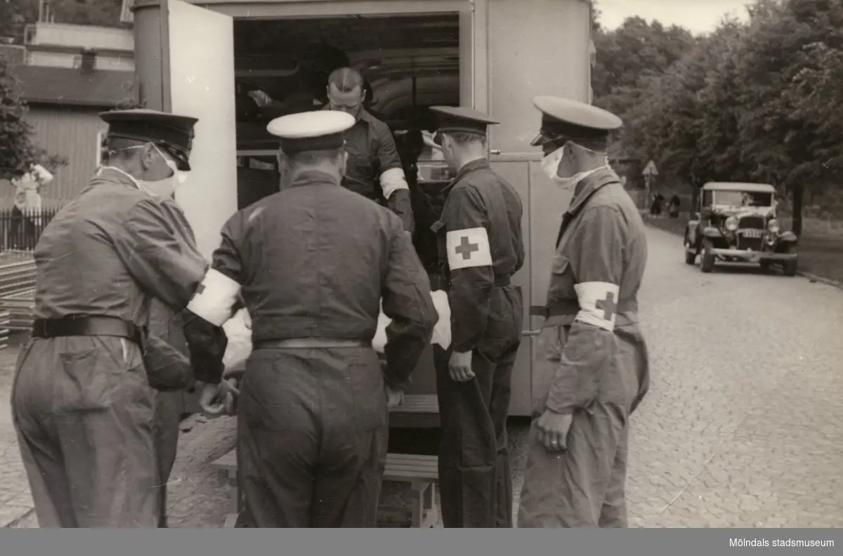 "Karantänsförläggning" på Kvarnbyskolan för överlevande från koncentrationsläger i Tyskland och Polen 1945.  
Skolan fungerade under denna sommar som beredsskapssjukhus.

Röda Korsets personal som hjälps åt att lyfta ut från buss eller ambulans, troligtvis en patient på bår.