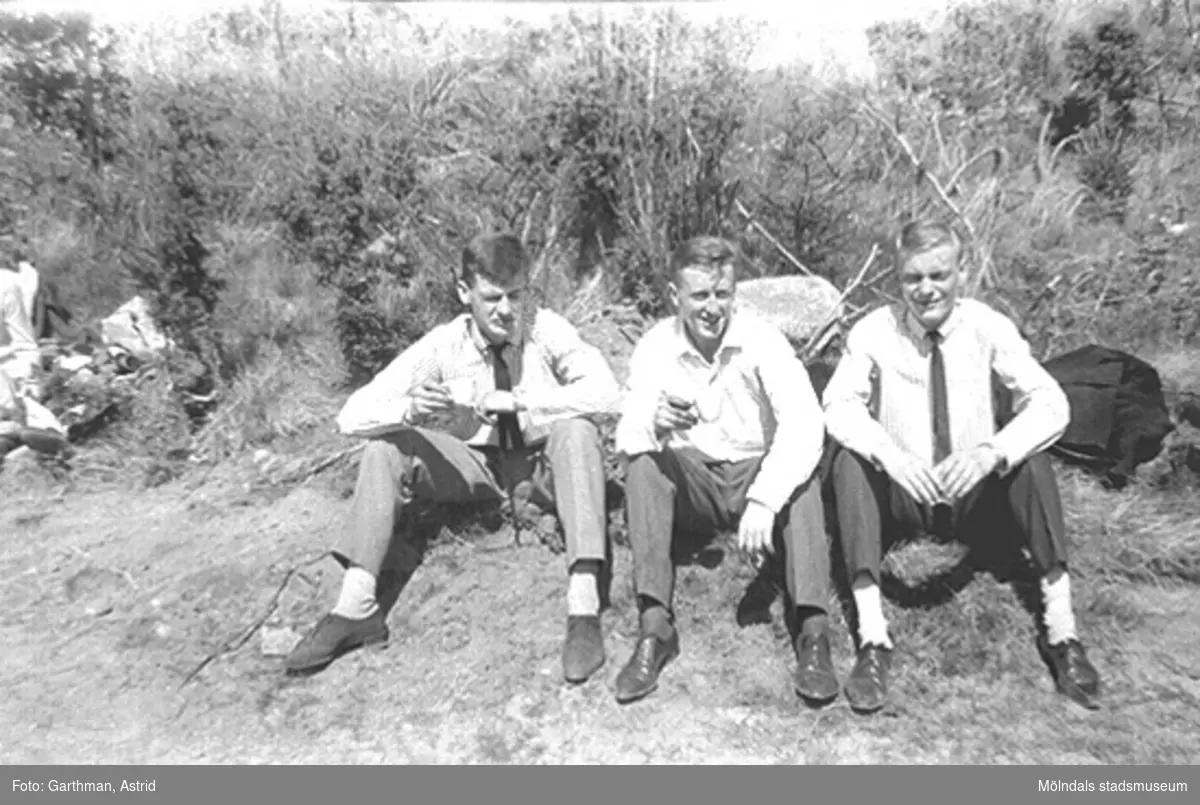 Söndagsutflykt till motokrossbanan i Blixås. Kamraterna Kjell Lager, Leif Garthman och Roger Karlsson sitter och fikar i solen. De unga männen är uppklädda, som man var på fritiden under 60-talet, i byxor, randig alt. vit skjorta, slips och lågskor. Blixåsbanan, 1965.
