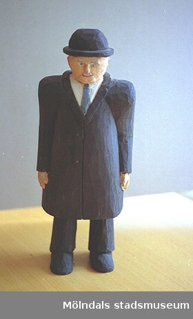 En gubbe står och tittar. Han är klädd i vit skjorta, svart rock, slips, hatt och byxor. Harry Bergmans "gubbar" (träfigurer).