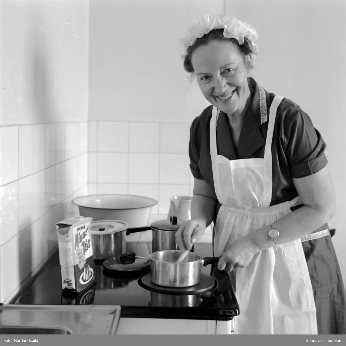 Kvinna i vitt förkläde och hätta fotograferad i köket och salongen. Svartvik.