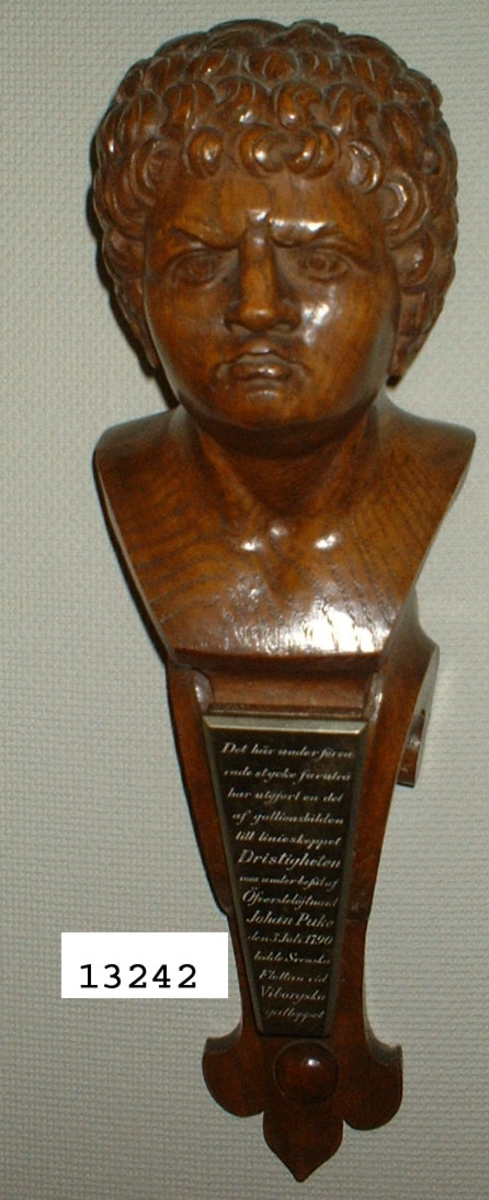 Galjonsbild, miniatyr. Föreställer linjeskeppet DRISTIGHETENS galjonsbild.
Text på mässingsplatta: År 1905 skänkte chefen för H.M. pansarbåten Dristigheten kommendörkapten Gustaf af Klint denna bild till fartygets gunrum.
Text på framsidan: Den här under förvarade stycke furuträ har utgjort en del av galjonsbilden till linjeskeppet Dristigheten som under befäl av överstelöjtnant Johan Puke den 3 juli 1970 ledde svenska flottan vid Viborgska viken.