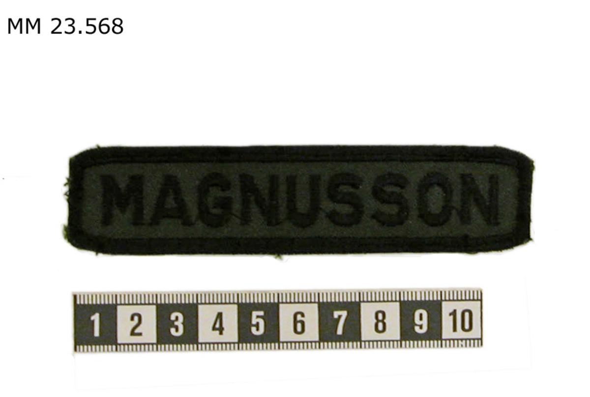Broderat namnband i svart text mot mörkgrön bakgrund. Ramen är svartbroderad. På namnbandet står "MAGNUSSON".