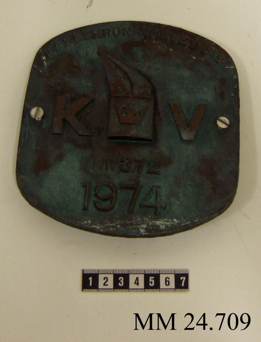 Metallskylt med inskription i relief. Texten på skylten lyder: Karlskronavarvet AB KV NR 372 1974. I mitten, dvs. texten KV, utformad som Karlskronavarvets logotype. Hål borrat i varje långsida.