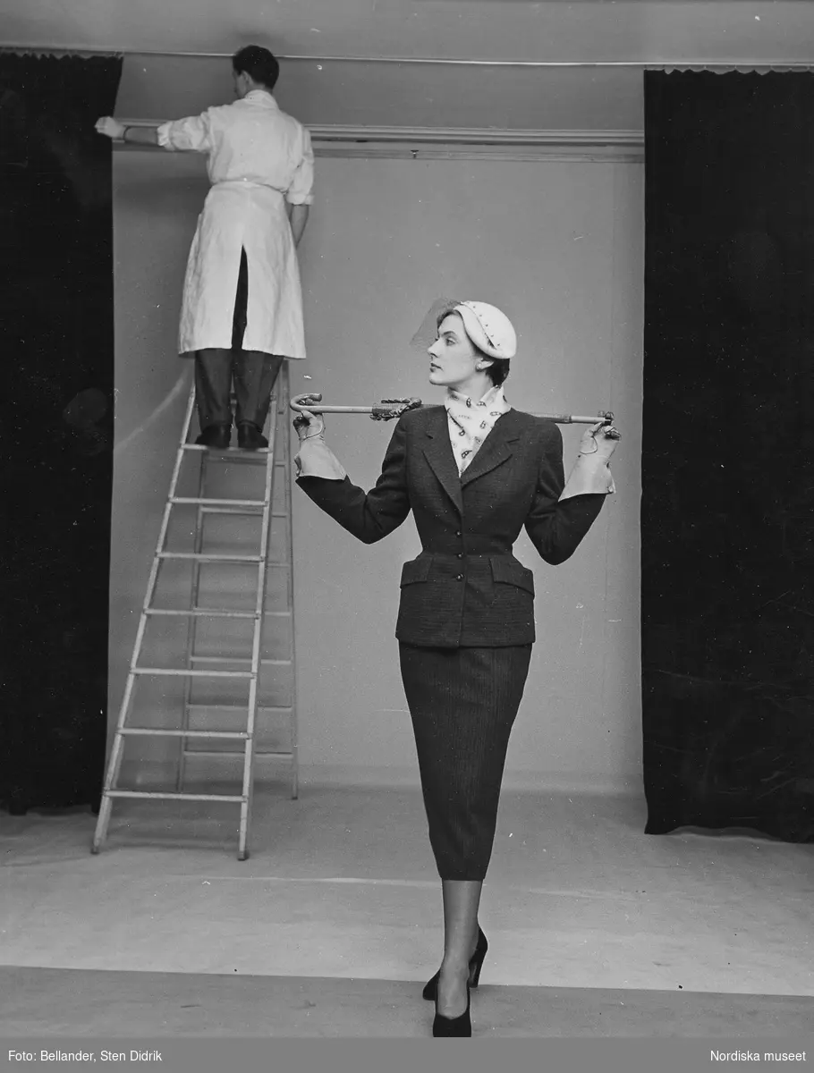 Mannekäng, troligen Ulla-Britt Enzell senare gift Gregemak (1953), klädd i dräkt, hatt med flor, scarf, handskar och pumps, håller i ett paraply. Man i vit rock i bakgrunden.