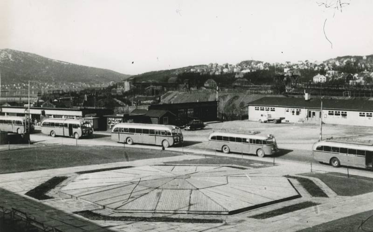 5 av Ofotens Bilruters busser passerer torvet i Narvik.  Fontenen er tildekker for vinteren.  Postbrakka til venstre..