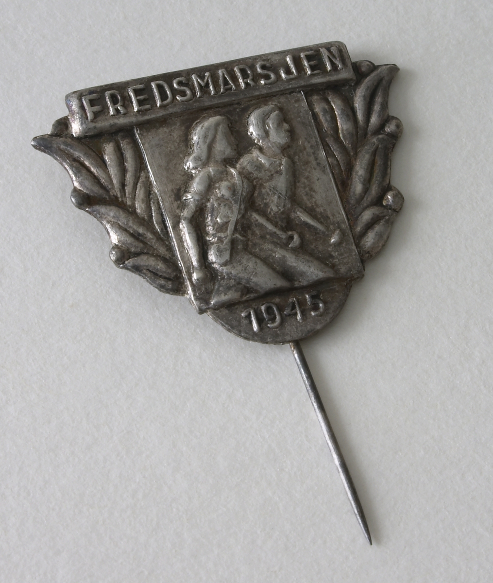 Nål laget av metall og har sølvfarge. På framsiden av nålen to personer med en bladranke på hver side. Inskripsjon: Fredsmarsen - 1945. Denne fikk Christine Knudsen, Foss for å gå Fredsmarsjen i 1945. 