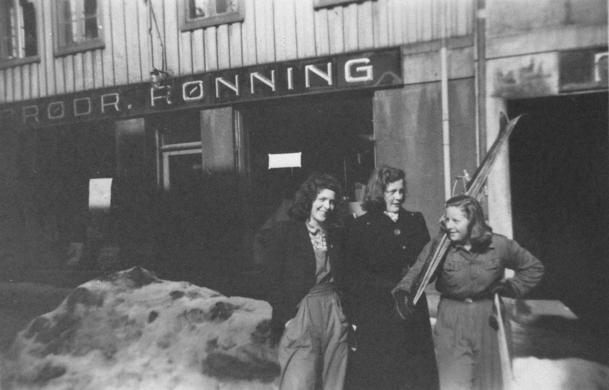 Tre unge kvinner utenfor bygning med skilt det står "Brødr. Rønning" på. Kvinnen til høyre bærer på ski og skistaver.