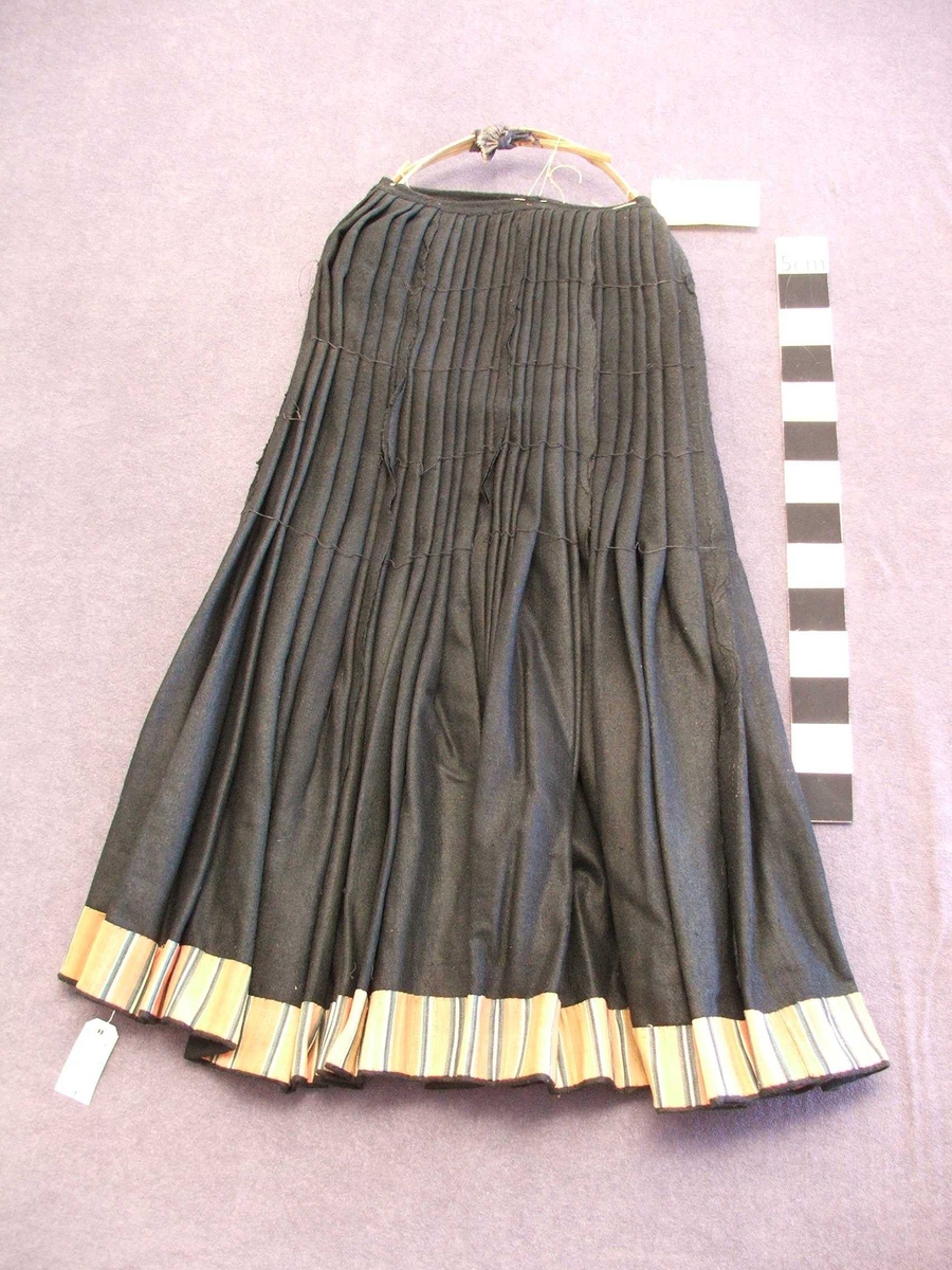 På vranga er det sydd kanting med 7,5 cm.bomullsstoff . Stoffet har loddrette striper i brunt, grønt, svart, raudt, blått.På ein del nedst: svart fløyelskant.