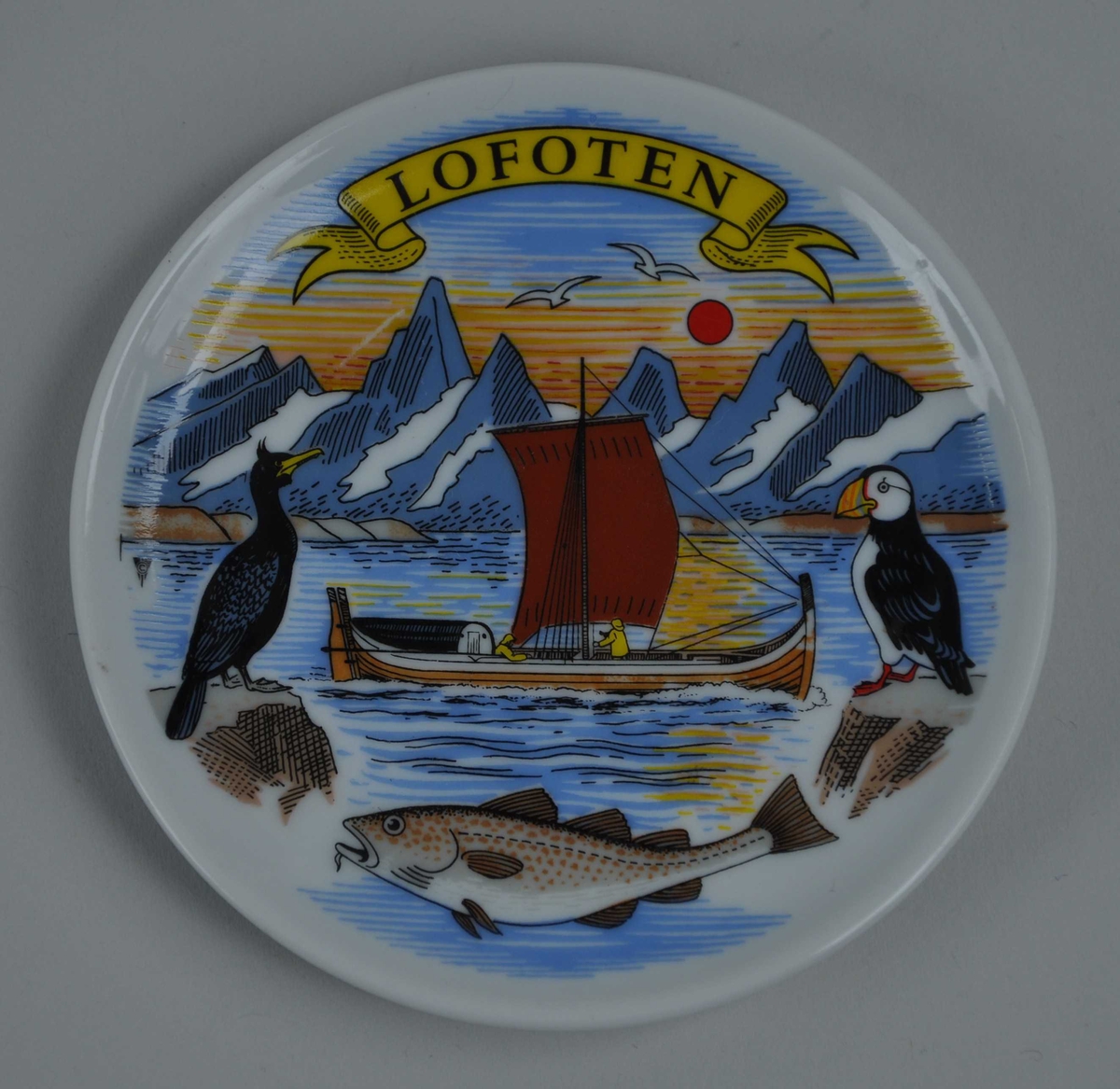 Motiv frå Lofoten, med Lofotveggen i bakgrunnen, ein Nordlandsbåt med segl og to fiskarar ombord, og i framgrunnen ein skarv, ein lundefugl og ein skrei. Lofoten står på eit gult banner øvst på brikka.