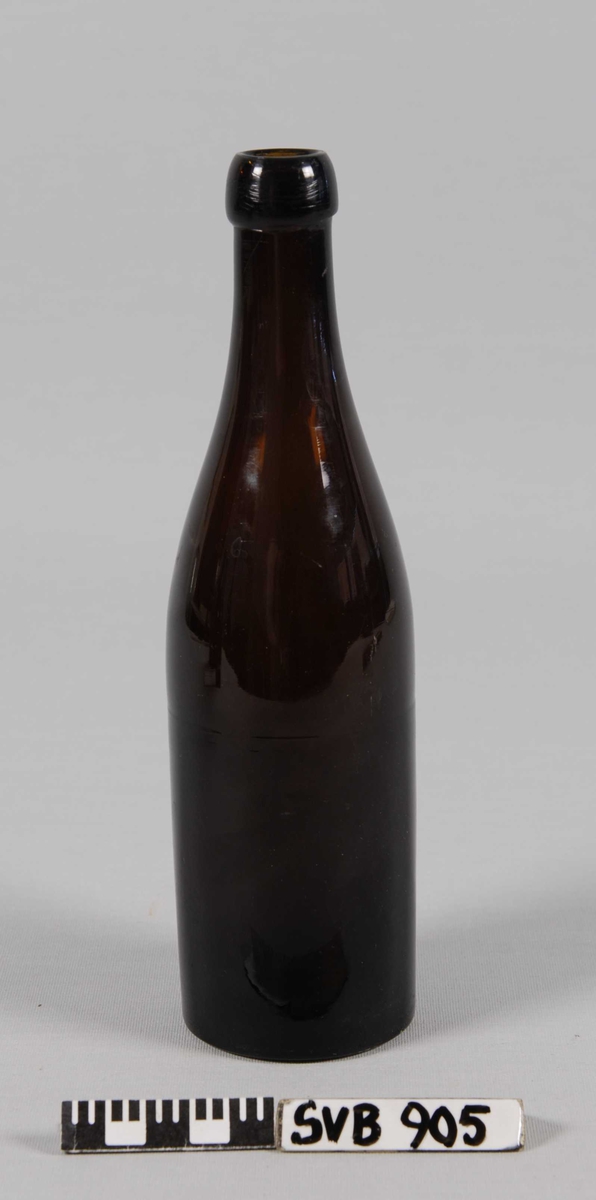 Drikkeflaske i brunt, klart glass. Slank kropp og lang hals med flat bunn. Klart avgrenset flasketut for propp av kork. 