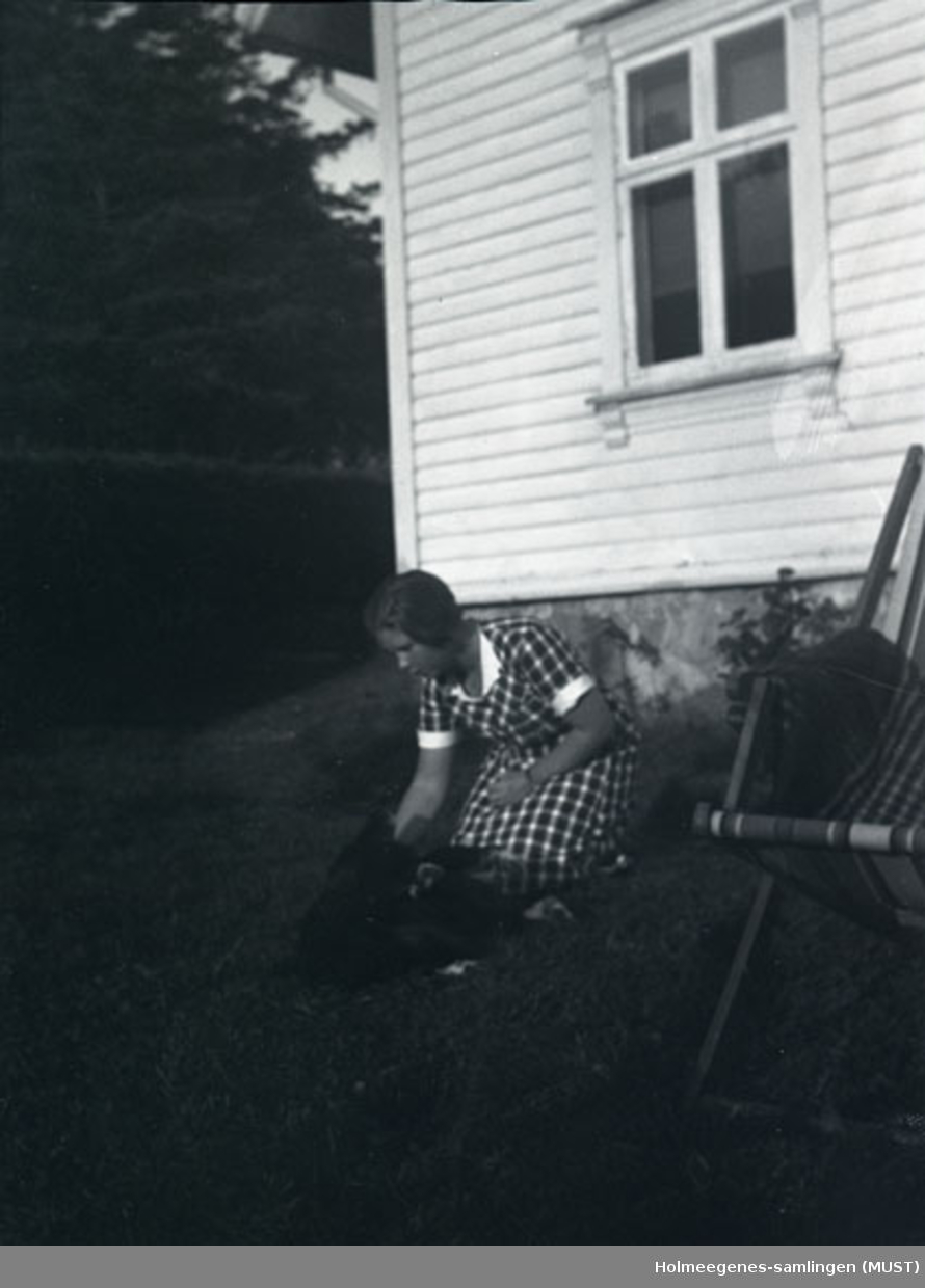 En ung kvinne i sommerkjole sitter på huk utenfor et hus. Det ser ut som hun klapper en hund (litt vanskelig å se pga. mørkt bilde). Antatt samme sted og tidspunkt på ST.K.HE 2010-011-0091 til -0094.
