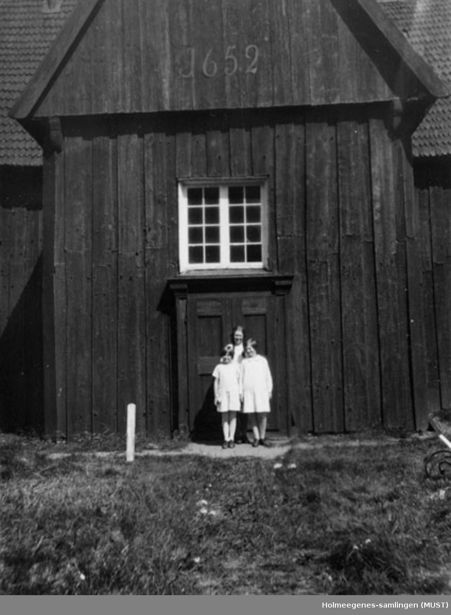 Tre jenter ved inngangen til en trekirke. Årstallet 1652 står over døren. Antatt Kvikne kirke på Tynset. Se ST.K.HE 2007-011-0125. Se også ST.K.HE 2007-011-0111 til -0126.