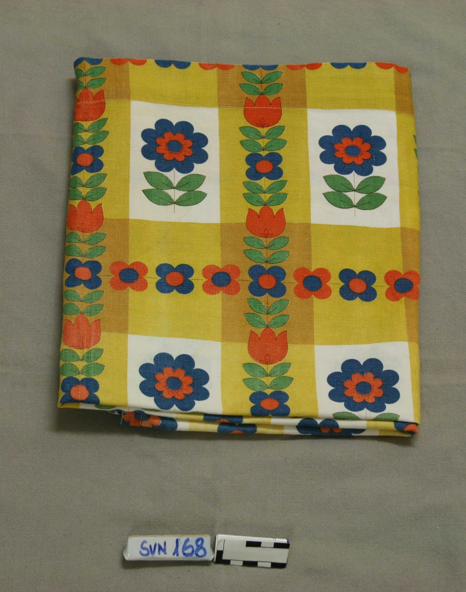 Rektangulær tekstilgardin. Mønster: hvite og lysebrune ruter med stiliserte røde, blå og grønne blomster.