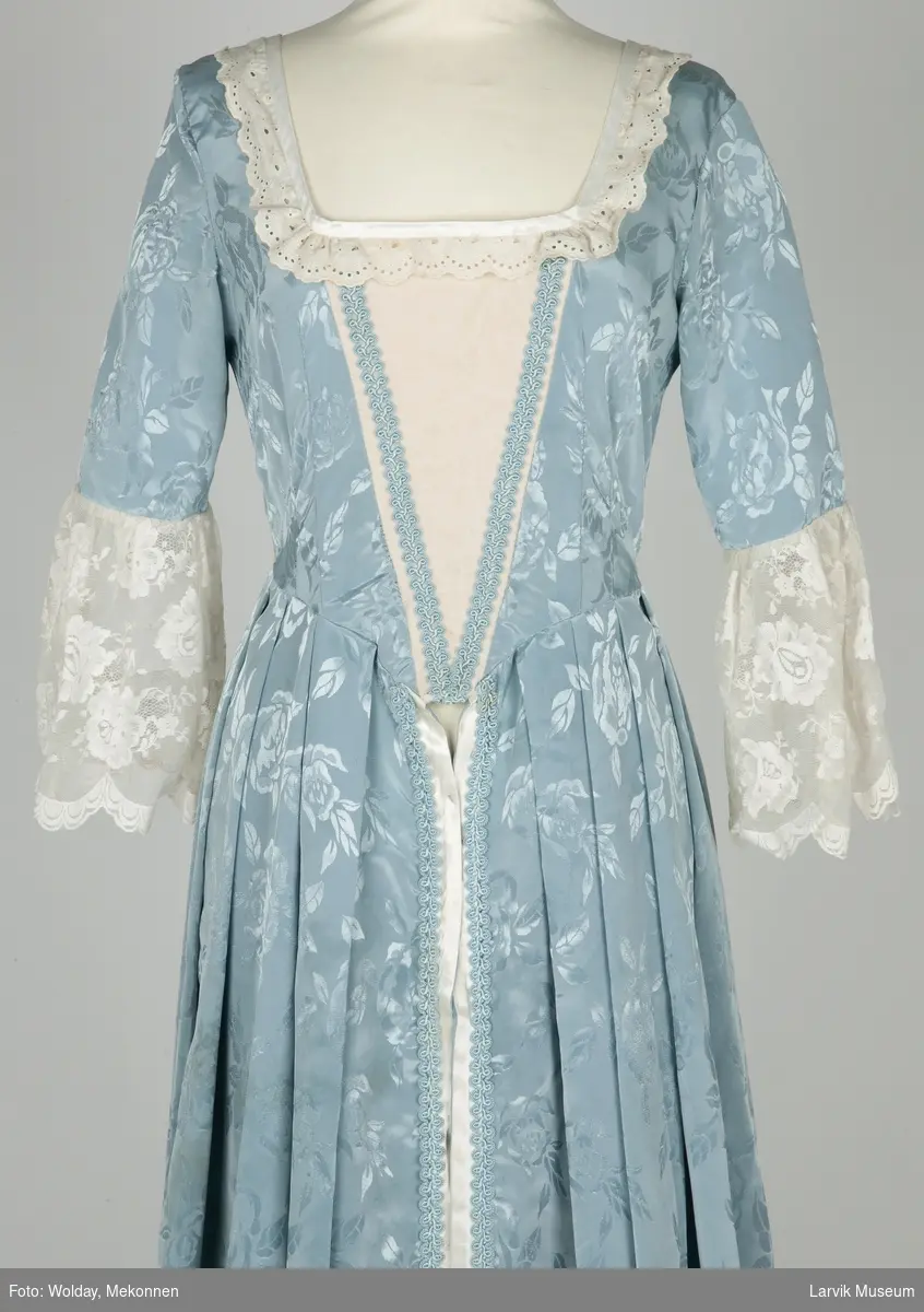 Lang kjole i lyseblå imitert silkeog blonder. Brukt som kjole/kappe under stivt underskjørt.