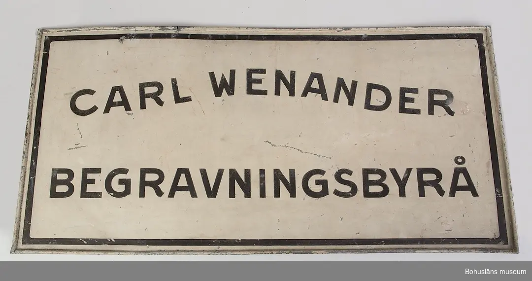 Vit plåtskylt med svart text och kant: 
CARL WENANDER
BEGRAVNINGSBYRÅ
Baksidan omålad. Troligen tillverkad 1900-1920.