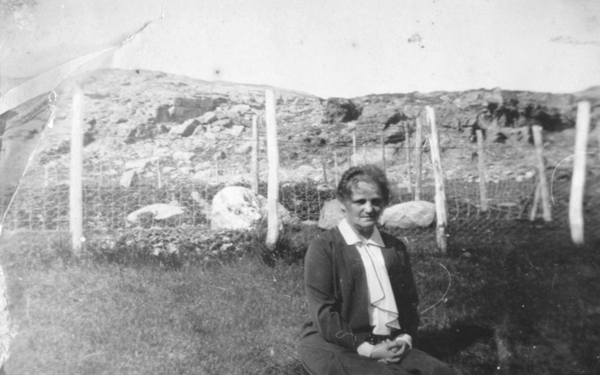 En kvinne sitter på bakken foran et nettinggjerde en sommerdag. Sted og person er ukjent, men bildet kan være tatt i Kvalsund kommune.