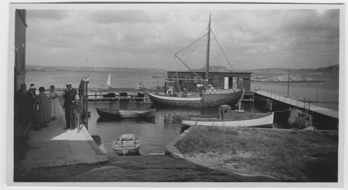 'Från Kristinebergs brygga. Magnus Aurivillius närmast. Vy mot havet med 6 personer på brygga intill byggnad. 2 av dem unga män med studentmössor. Liten hamn med fiskebåtar och ekor. ::  :: Serie fotonr 1888-1963.'