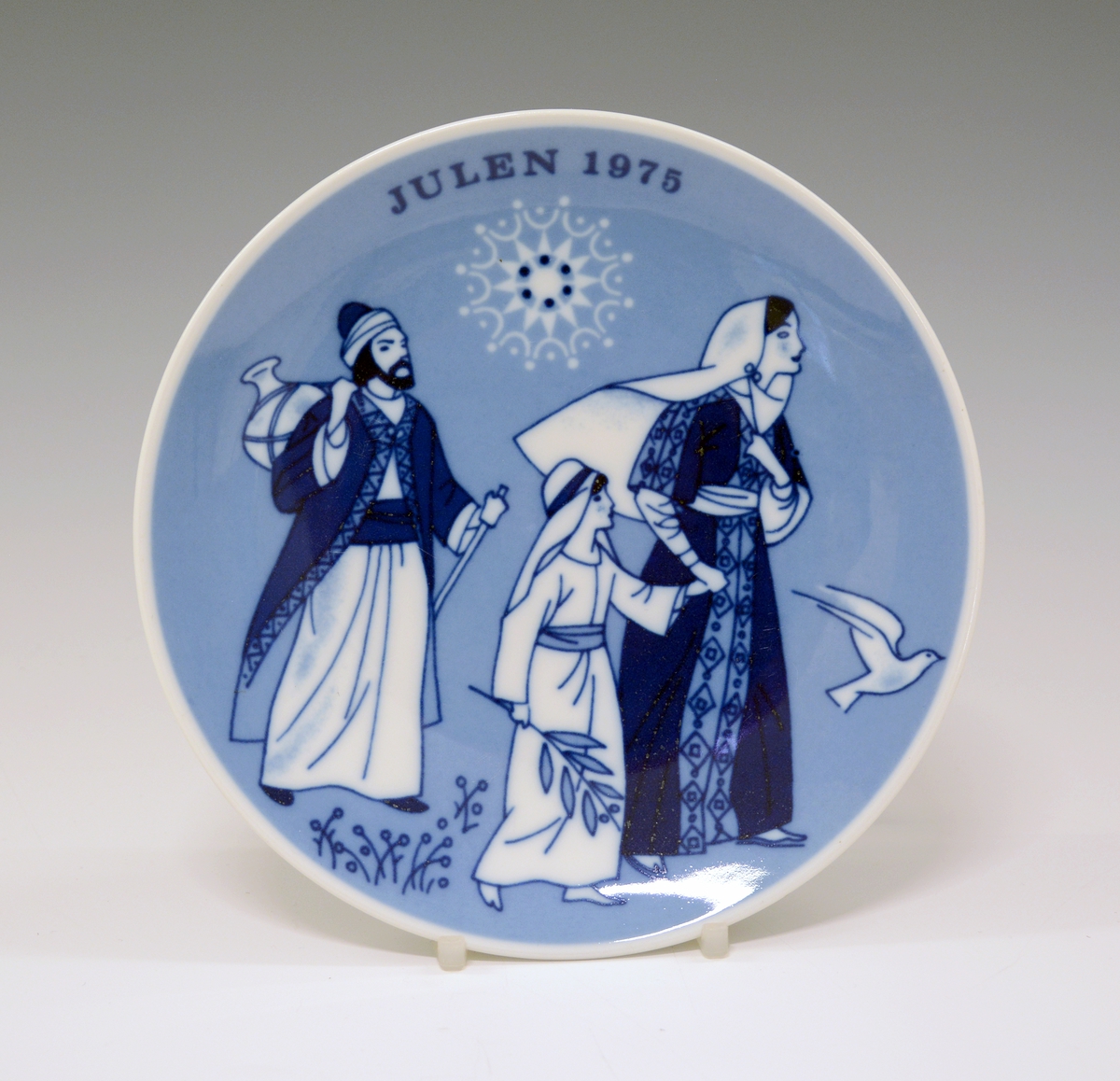 Juleplatte av porselen. Glatt modell. Hvit glasur. Dekorert med Jesus, Maria og tekst Julen 1975 i ulike blåfarger.
Dekor av Gunnar Bratlie.