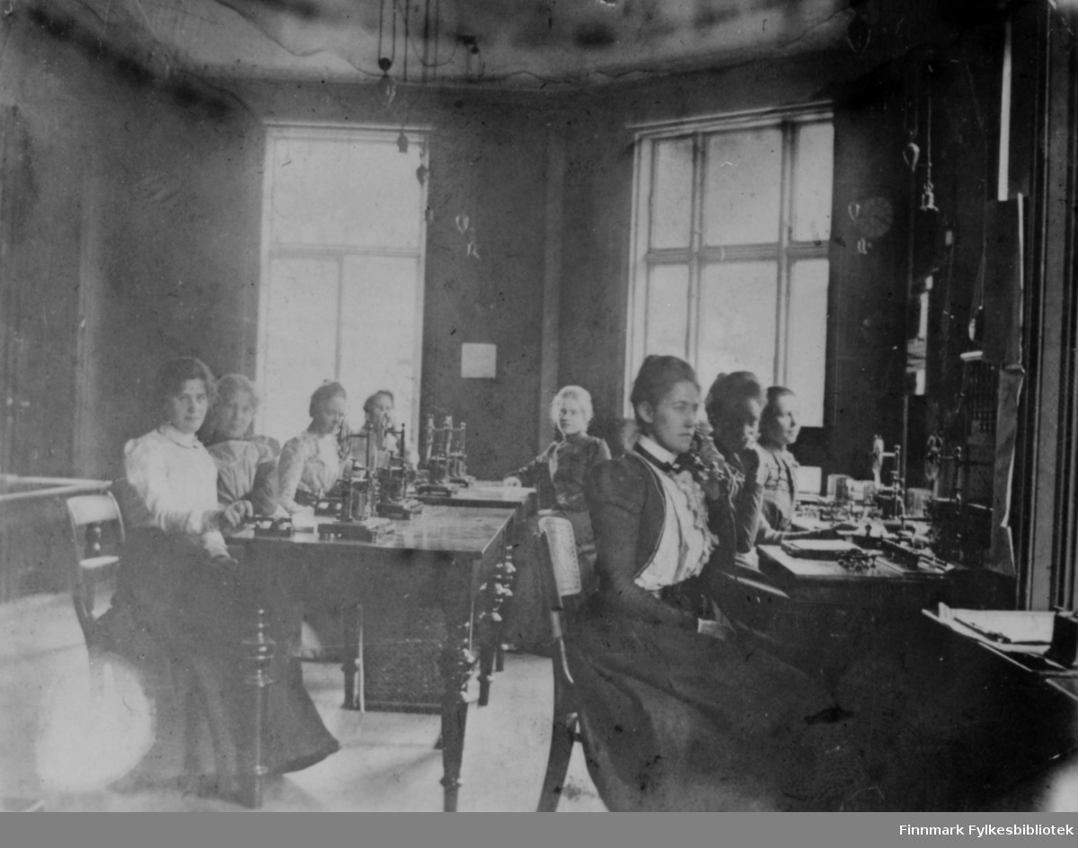 Åtte kvinner sitter ved arbeidsbordene sine ved telegrafekspedisjonen i Hammerfest. Alle er kledd i lange kjoler og har telegrafsender foran seg, også kalt morsenøkkel eller telegrafnøkkel.
