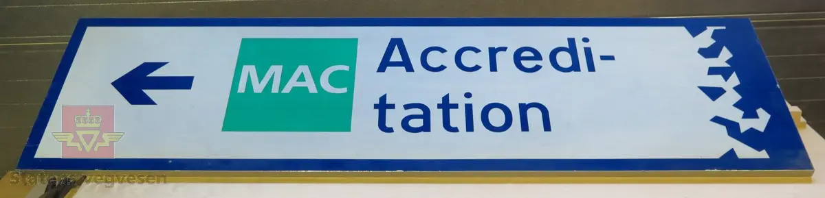 Rektangulært skilt av aluminium. Har symbol for kjøreretning samt teksten "MAC Accreditation". Blått, grønt og hvitt, med grå bakside.