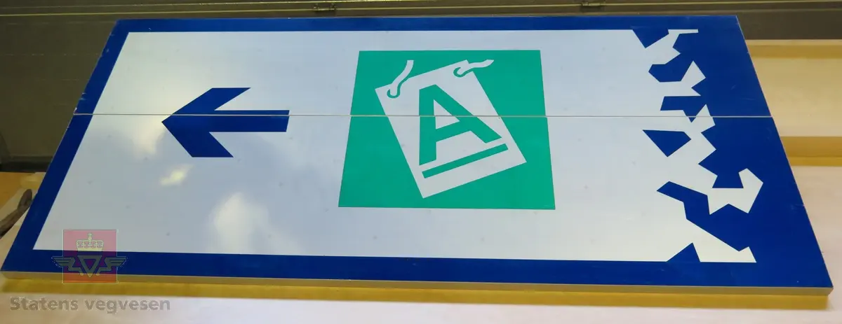 Rektangulært skilt av aluminium. Har symbol for kjøreretning samt teksten "A". Flerfarget, med grå bakside.
