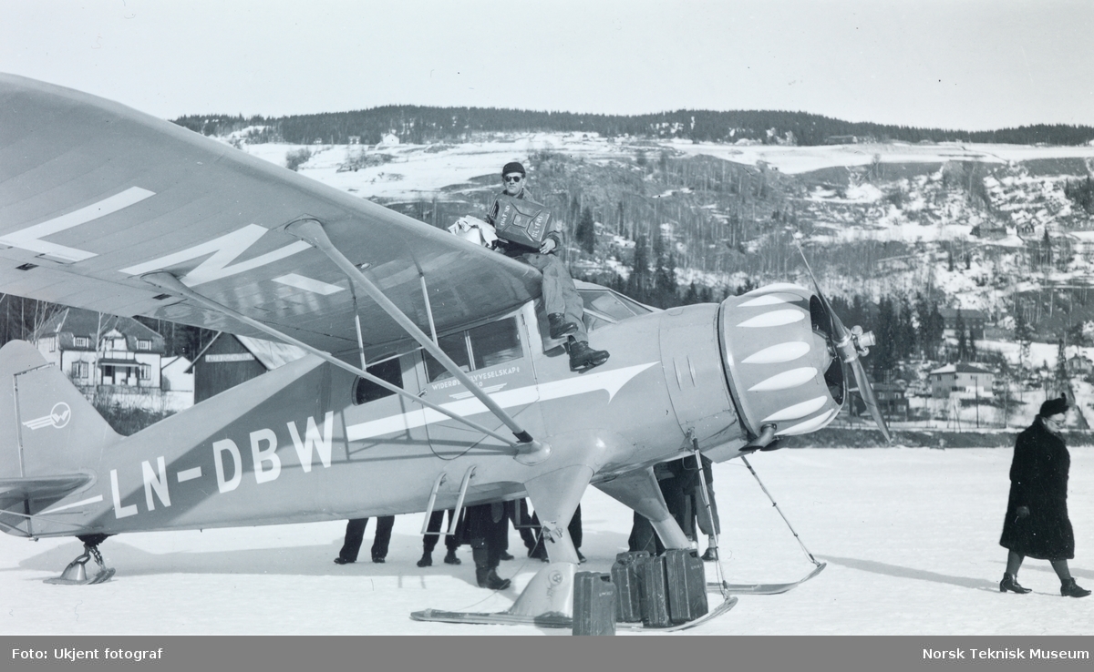 Flyet "Hønningstad C-5 Polar" står på et snødekket vann. En mann sitter på toppen av flyet og fyller drivstoff.  Flyet er merket med Widerøe Flyveselskap og registeringsnummer "LN-DB W"