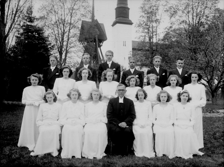 20 konfirmander, 14 flickor, 6 pojkar och kyrkoherde Johansson.
Glanshammars kyrka och klockstapeln i bakgrunden.