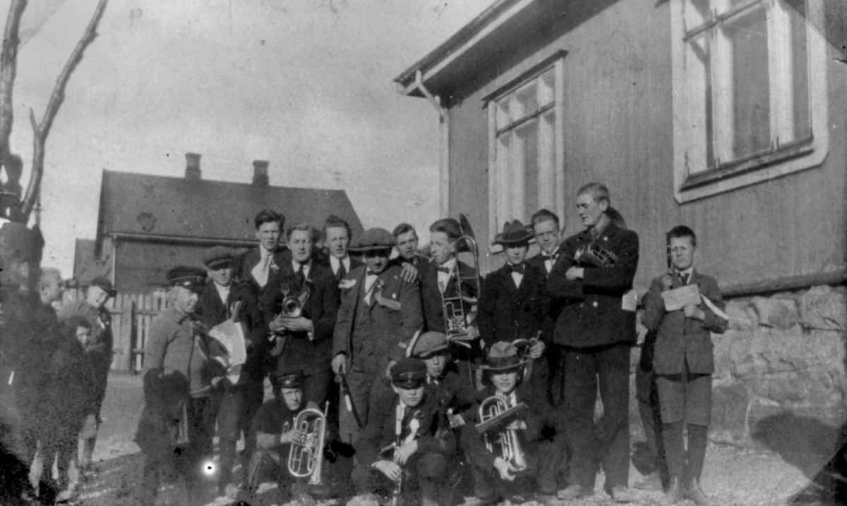 Kirkenes hornmusikk. 15 gutter og menn med instrumenter. Foto- grafert utendørs. Et par hus i bakgrunnen. Ingen årstall.