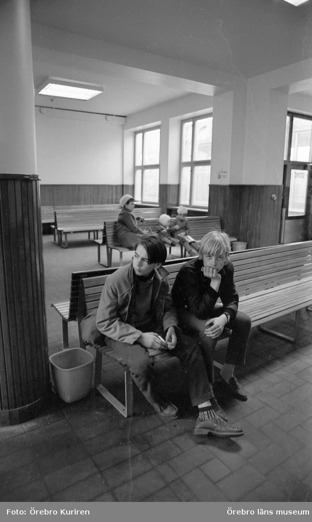 Hallsberg 10 maj 1969

I en väntsal sitter två pojkar på en träsoffa. Det sitter också en kvinna i kappa och mössa med två barn.