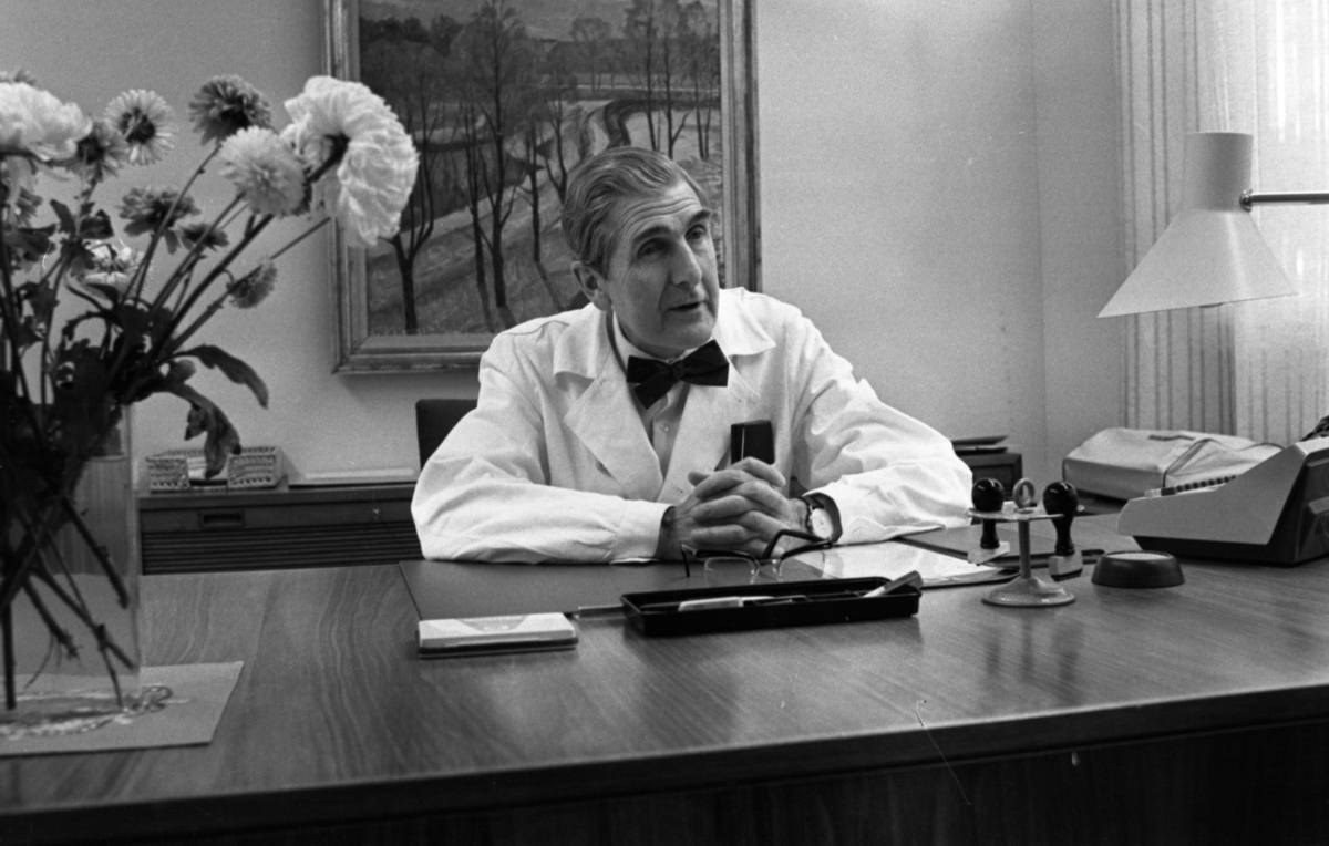 Lungkliniken, Nappivalen, Grekiskt handarbete 11 december 1967

En läkare på lungkliniken sitter vid bordet inne på sitt kontor. Han är klädd i en vit läkarrock, vit skjorta och svart fluga. En blomvas står med en blomma till vänster i på en liten duk på bordet. Bakom doktorn hänger en tavla på väggen.