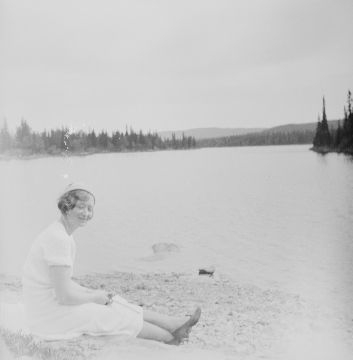 En kvinne sitter på en steinete strand ved en innsjø i Jeppedalen. I bakgrunnen ligger skogkledte nes og åser.