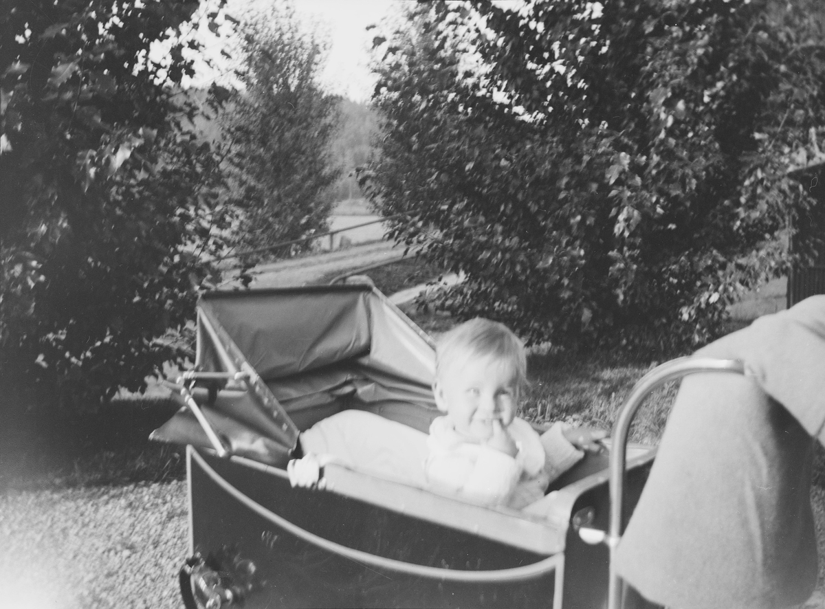 Et lite barn, lille Pierre, sitter i en barnevogn som står på gårdsplassen til Linderud Gård ved sommerstid. Barnet smiler og "tygger" på en finger.
