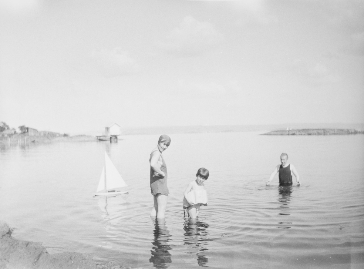 Celina Marie, et barn og Iacob Ihlen Mathiesen bader i sjøen. En liten seilbåt, en leke, flyer godt på stille vann. En brygge med et lite hus ytterst sees i bakgrunnen.