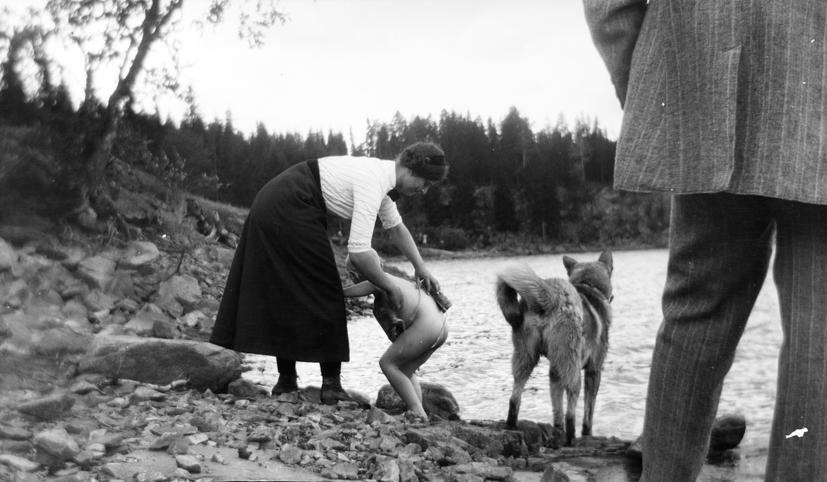 En kvinne hjelper et barn med håndkle og tørking etter et bad i innsjøen. En elghund står ved siden av dem i strandkanten. Foran står en mann med ryggen til kameraet. Bildet er kanskje tatt i Jeppedalen.