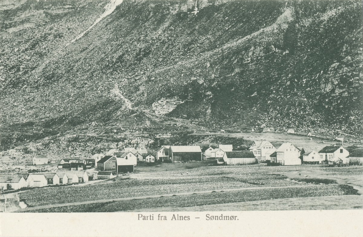 Prospektkort flere gårdstun og naust på Alnes, Godøy. I fjellterrenget bak ses stien opp til fjellet.