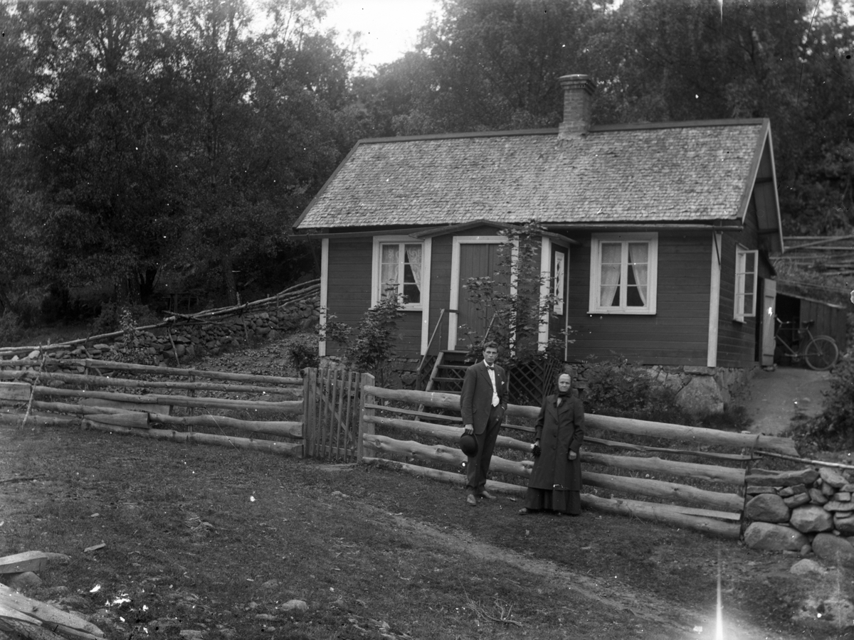 Mor och son fotograferad utanför torp med staket av trästockar. Torpet låg på Stackenäs gårds ägor.
