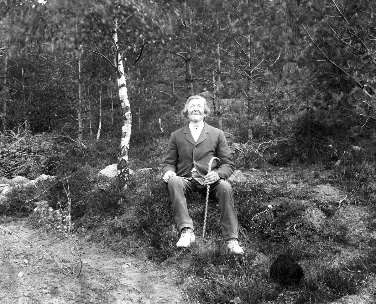 En man, Abraham Johansson, sitter på en sten vid vägkanten med skog i bakgrunden. Han håller en käpp i handen.