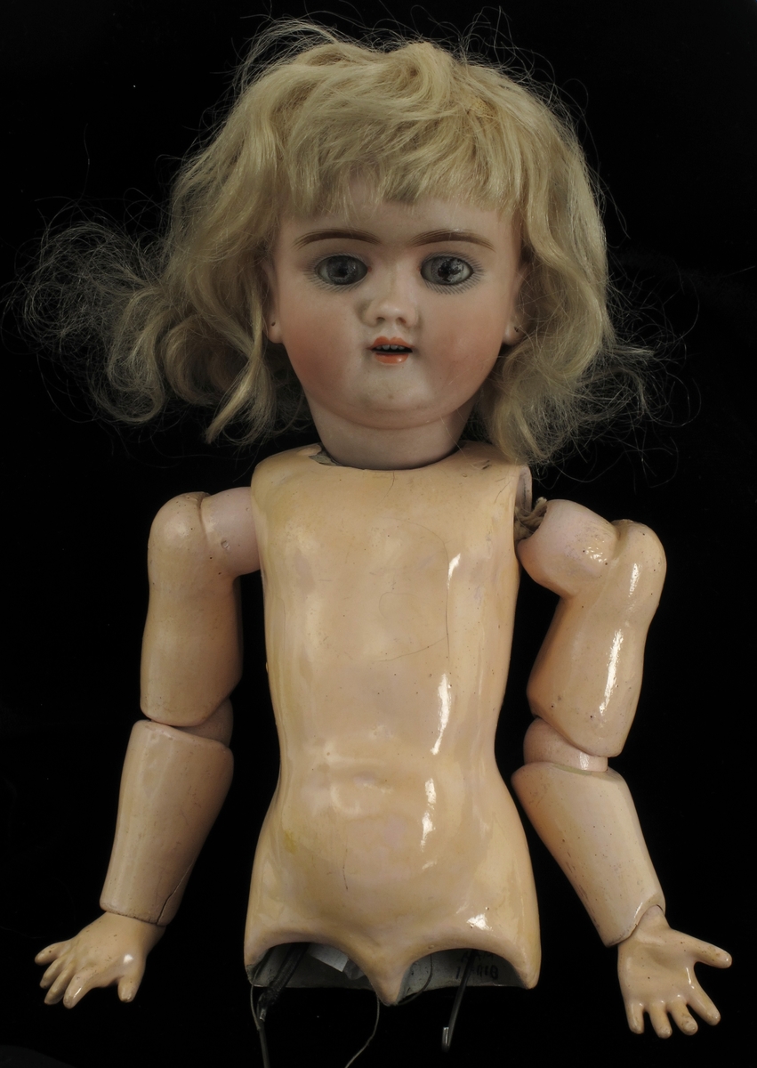 Dukke. Hode av porselen, med øyne som lukkes når dukken ligger, med lyst hår. Kropp av pappmasjé  påtrykt  Handwerek".